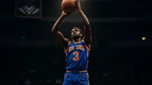 Knicks player shooting three-pointer at Madison Square Garden with championship banners visible