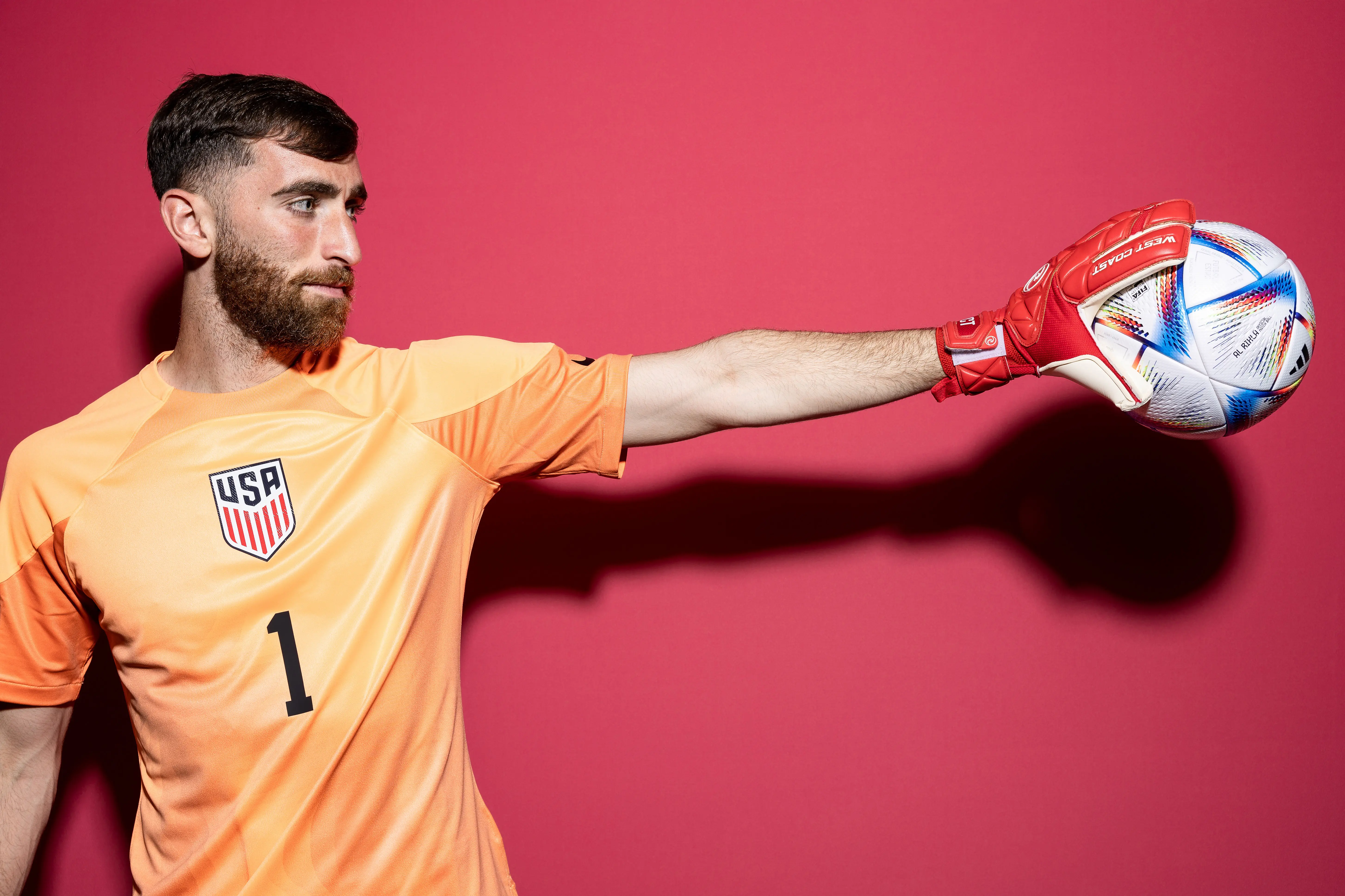 USMNT goalkeeper in orange jersey holding a soccer ball against a pink background.