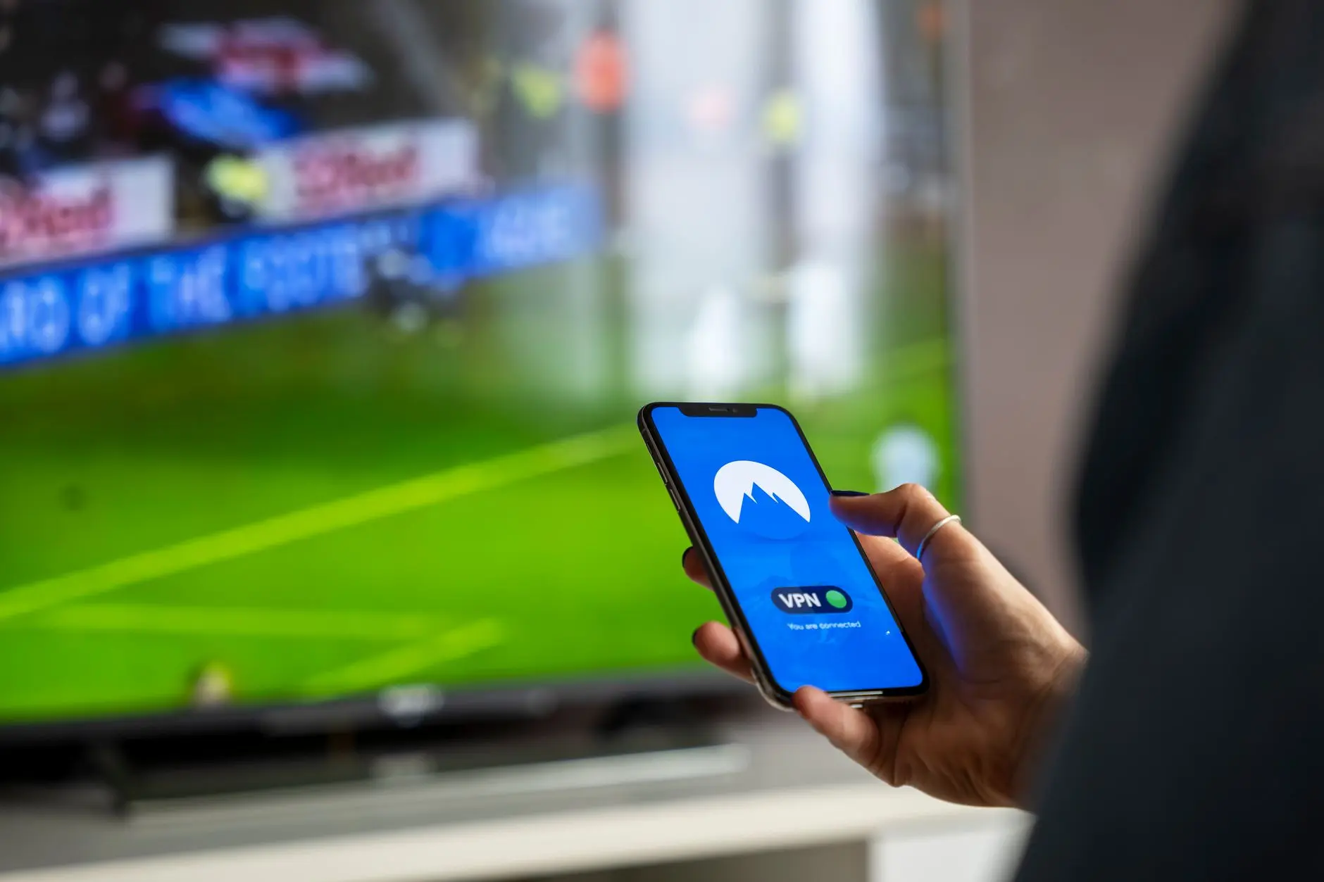 A person holding a smartphone displaying a VPN app while watching a sports event on TV.